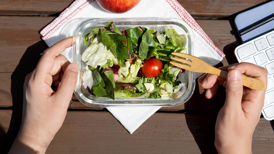 Brown table with disposable lunch box filled with salad, apple and napkin, two hands near the lunch box of a person ready to eat