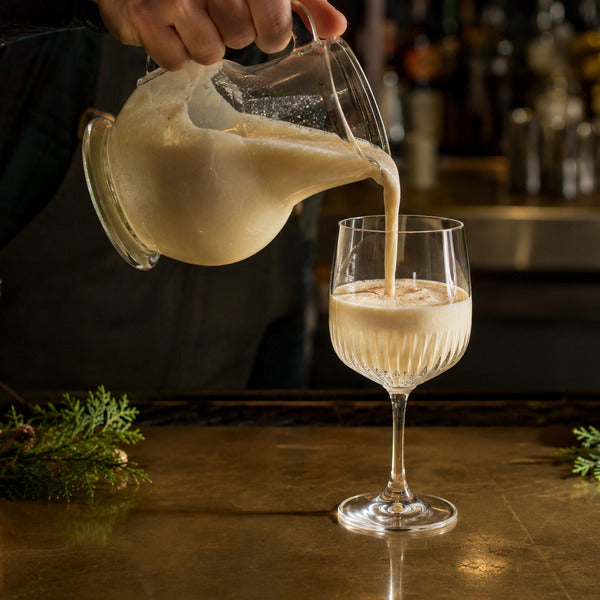 Bartender pouring classic eggnog from a glass pitcher into a Voglia Nude crystal water goblet on a holiday-decorated bar