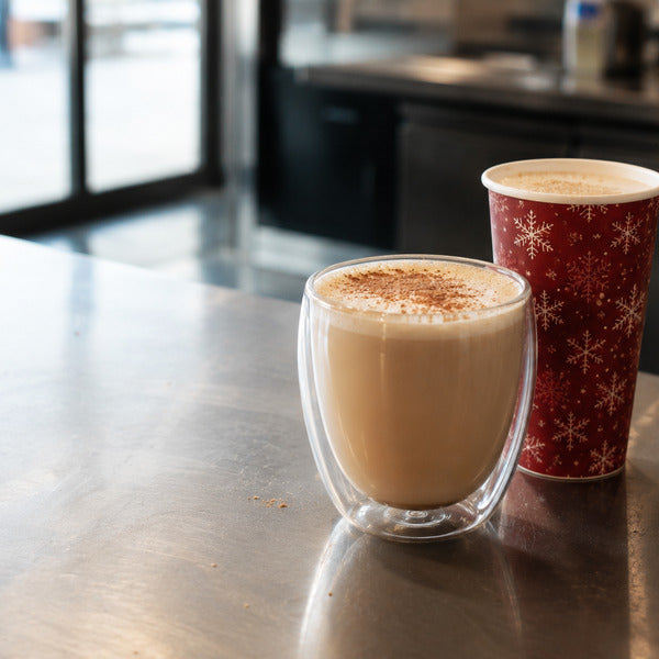 Eggnog latte served in a Forma Professional double wall glass beside a Restpresso red Christmas snowflakes paper to-go cup on a cafe counter