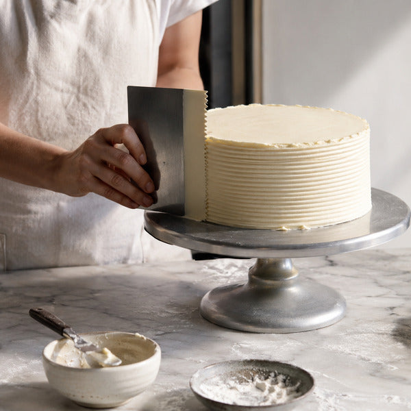 Baker smoothing buttercream cake frosting on a revolving turntable using a metal cake scraper and icing comb for a striped finish