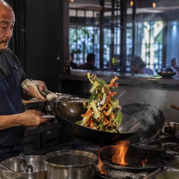 Chef tossing vegetables in a seasoned carbon steel wok over a high-heat commercial burner