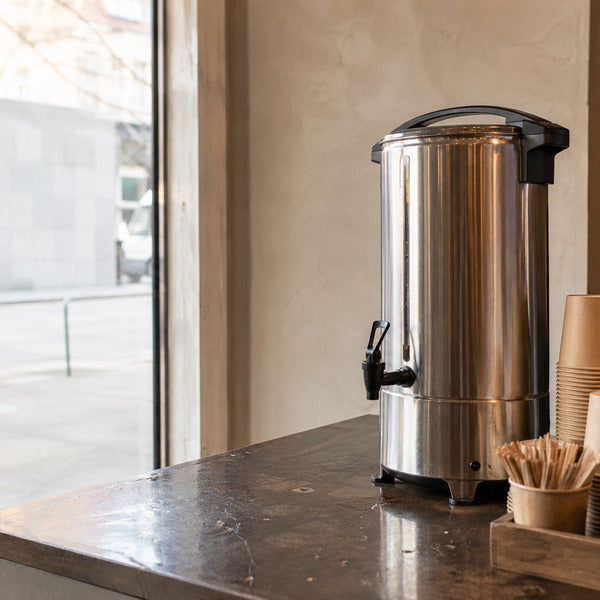 Large coffee urn dispenser set up for a catering event with cups, stirrers, and creamer nearby