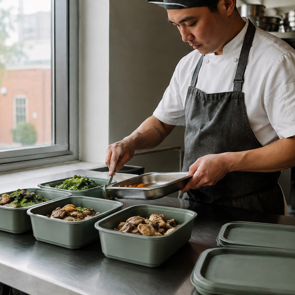 Chef filling commercial-grade reusable food containers on a restaurant prep line for a takeout program