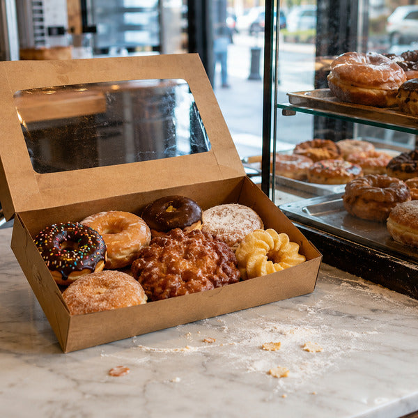 Assorted types of donuts packed in a windowed kraft bakery box beside a bakery display case