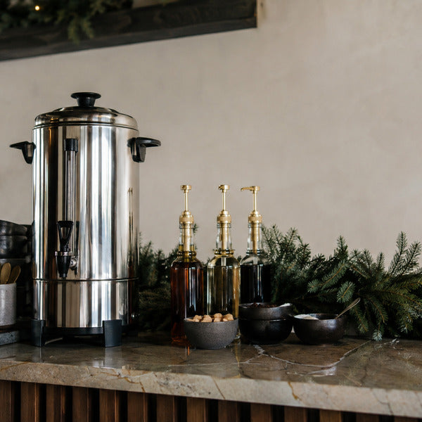 Festive holiday coffee bar ideas: garland-trimmed station with coffee urn, mugs, and syrup bottles