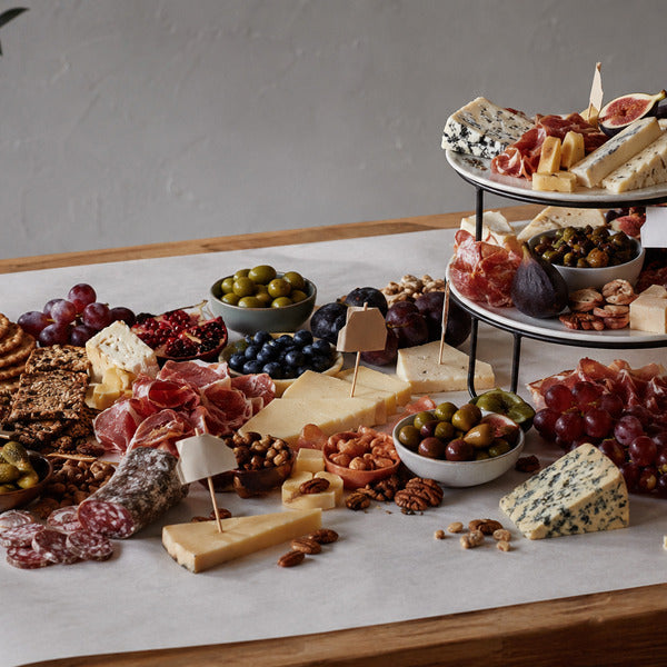 Grazing table spread with a tiered platter of cheeses and cured meats, plus grapes, olives, pomegranate, mixed nuts, and crackers arranged across the table