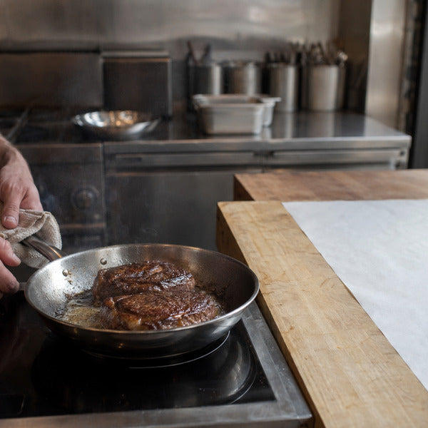 Line cook searing steaks in a stainless steel fry pan on an induction range in a commercial kitchen