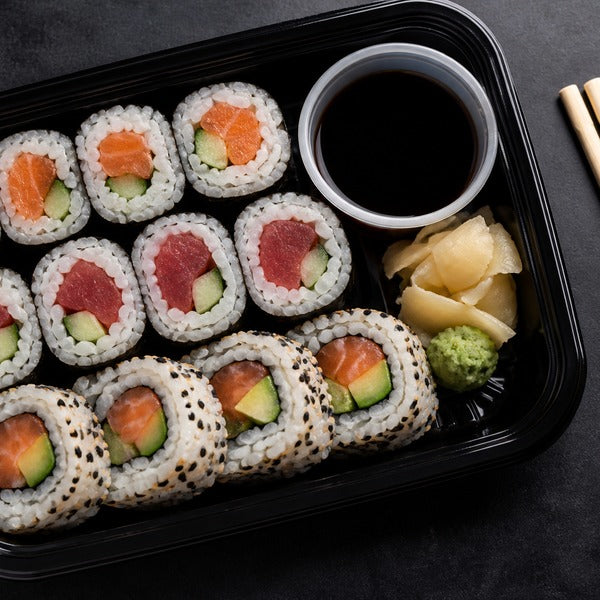 Takeout sushi rolls packed in a black plastic sushi container with clear lid, soy sauce cup, and wrapped bamboo chopsticks