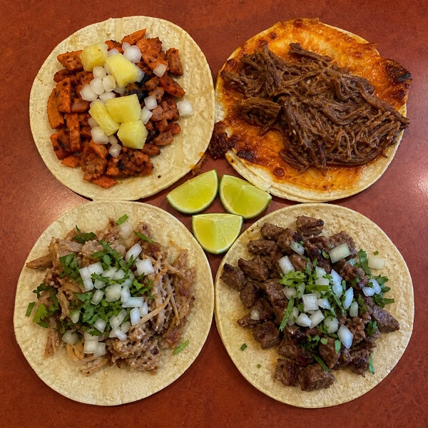 Four types of tacos side by side — tacos al pastor, birria tacos, carnitas, and carne asada on corn tortillas
