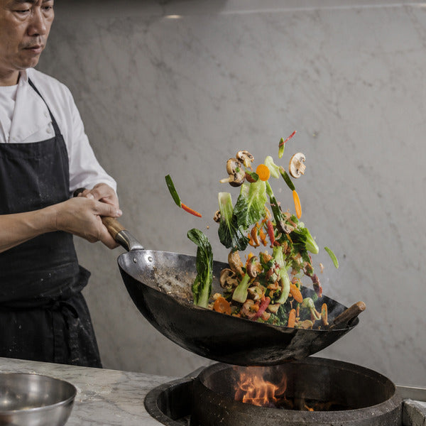 Chef tossing vegetables in a carbon steel wok pan over a high-heat commercial burner